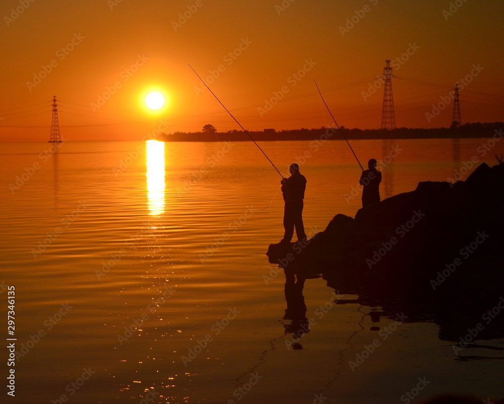 silhouette of two fisherman fishing on the background of the setting sun by the river