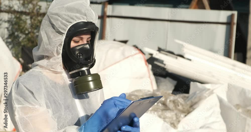 Woman in rubber gloves, military gas mask and chemical protection suit is standing in landfill in city limits, writes on paper violations found, makes inspection of garbage, draws up protocol Woman in rubber gloves, military gas mask and chemical protection suit is standing in landfill in city limits, writes on paper violations found, makes inspection of garbage, draws up protocol