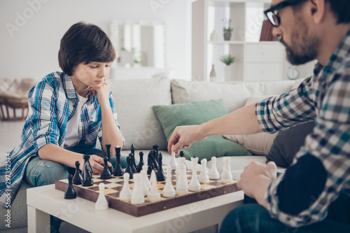 Portrait of two nice skilled focused serious guys dad and pre-teen son sitting on sofa playing chess moving pieces thinking strategy in light white modern interior house living-room