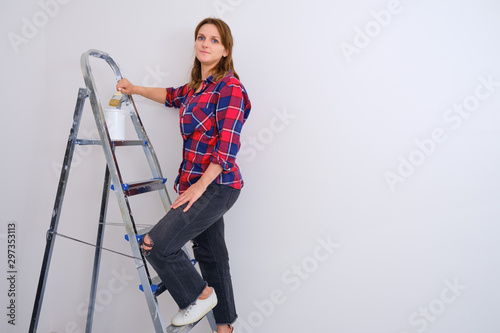 Woman painter stands on a stepladder in a plaid shirt, copy space. Painting the walls in a new apartment
