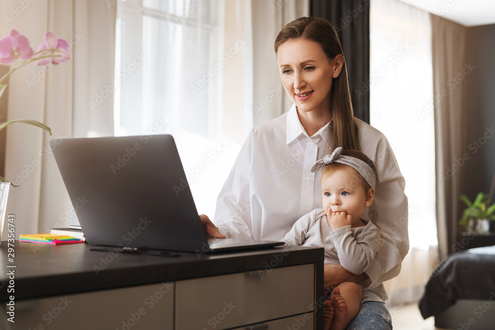 Female freelance author typing on laptop and holding baby on knees. Vacancies for home-based remote work for young parents. Distant career for professionals in digital segment with little children. Female freelance author typing on laptop and holding baby on knees. Vacancies for home-based remote work for young parents. Distant career for professionals in digital segment with little children.