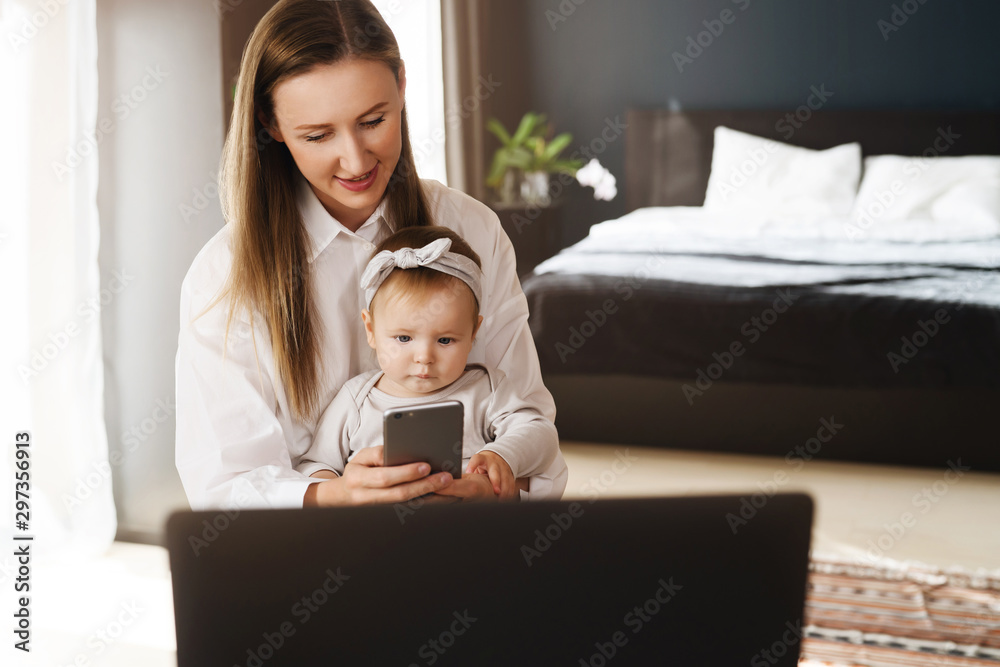 Young mother with daughter sitting in bedroom and enjoying watching interesting cartoon on smartphone. Ways to entertain child with online content and educating lessons for infants on digital gadgets. Young mother with daughter sitting in bedroom and enjoying watching interesting cartoon on smartphone. Ways to entertain child with online content and educating lessons for infants on digital gadgets.