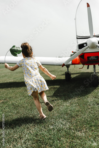Little girl with brown hair running on the green grass near airplane. Cute girl in dress posing on the aircraft. Walk at airport
