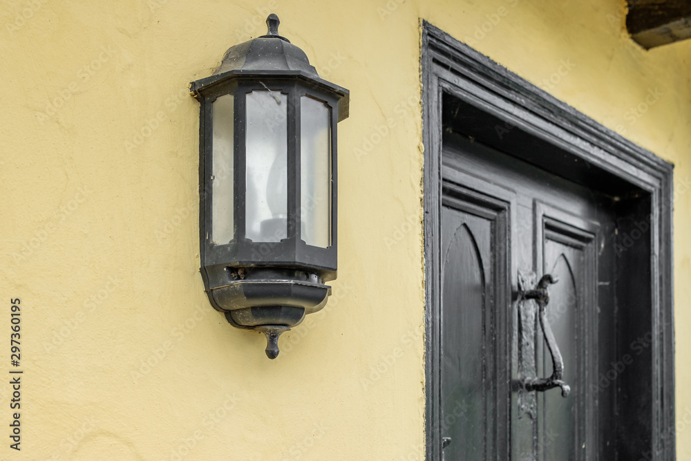Detailed, shallow focus of a wrought iron door lamp seen at the front of a very old, historic house. The wooden door and antique door knocker are also visible.