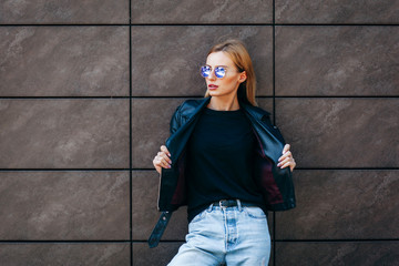 Girl wearing t-shirt, glasses and leather jacket posing against street , urban clothing style. Street photography