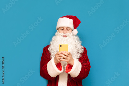 Smiling young man in santa suit standing on blue background and using smartphone, isolated. Santa teenager with smartphone isolated on blue background. Christmas concept.