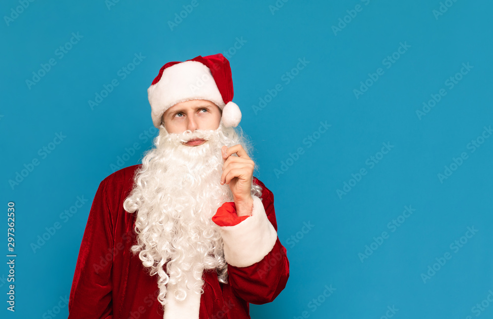 Closeup portrait of a pensive santa on a blue background, looking away at an empty space and thinking. Young guy in santa suit isolated on blue background. christmas concept.