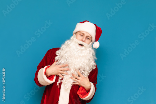 Positive young man in Santa suit standing on blue background, smiling and looking at camera. Young cheerful Santa laughing, isolated. Christmas concept.