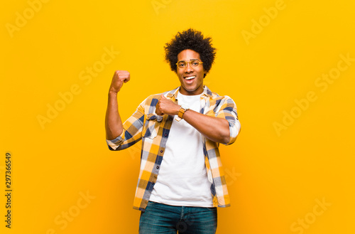 young black man smiling cheerfully and casually pointing to copy space on the side, feeling happy and satisfied against orange wall