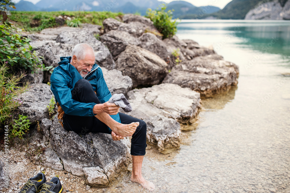 A senior man hiker sitting by lake in nature, taking shoes off.