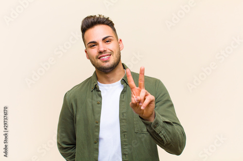 young hispanic man smiling and looking happy, carefree and positive, gesturing victory or peace with one hand against isolated background photo