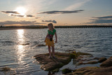 Young girl stood on rock at edge of water with sunset behind her