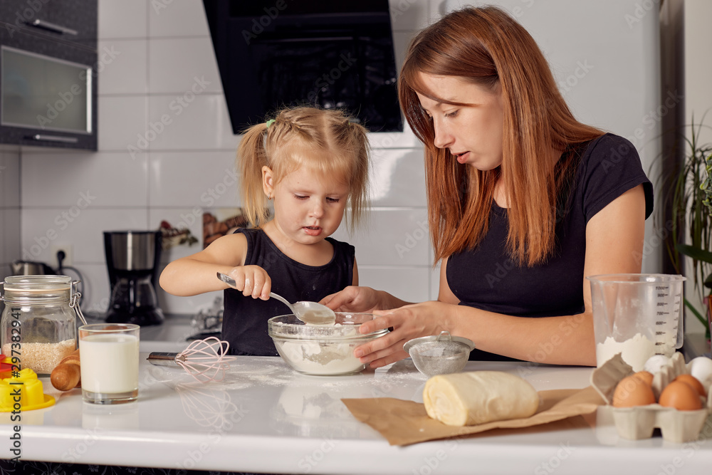 Mother and daughter preparing a sweet cake using flour, milk, sitting on chairs at a table in a modern kitchen. Girl holding a whisk, stirring eggs in a bowl, preparing pancake dough with her mom. Mother and daughter preparing a sweet cake using flour, milk, sitting on chairs at a table in a modern kitchen. Girl holding a whisk, stirring eggs in a bowl, preparing pancake dough with her mom.