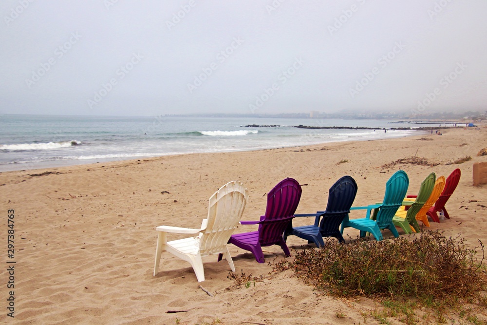 Colored Adirondack chairs face the Pacific Ocean on a sandy beach. Colored Adirondack chairs face the Pacific Ocean on a sandy beach.
