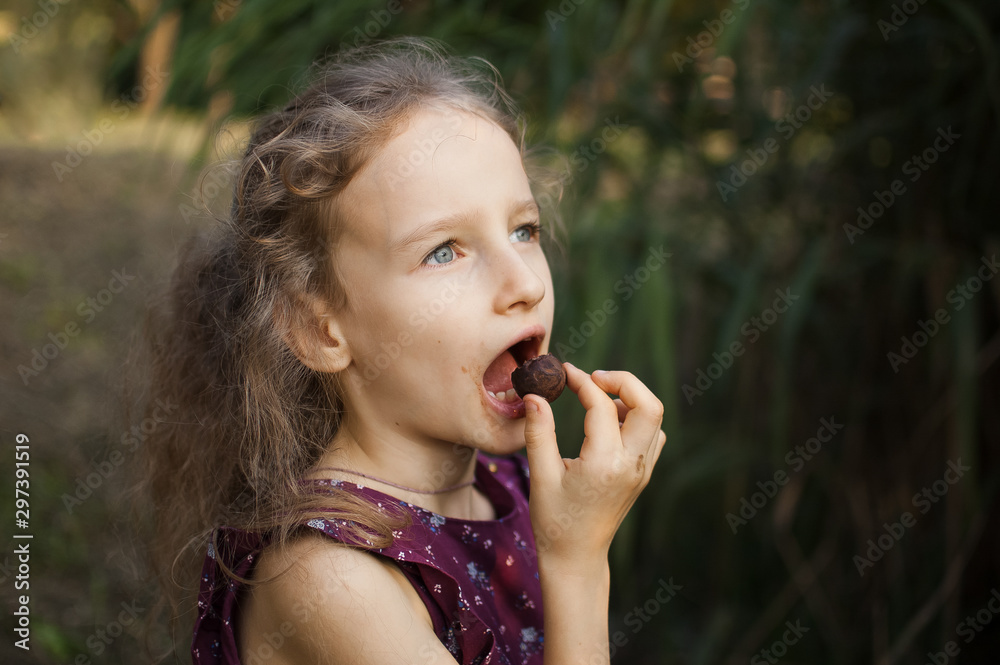 Beautiful little blond girl in violet dress with ponytail is eating a chocolate candy in the park durring a day, sugar addiction concept