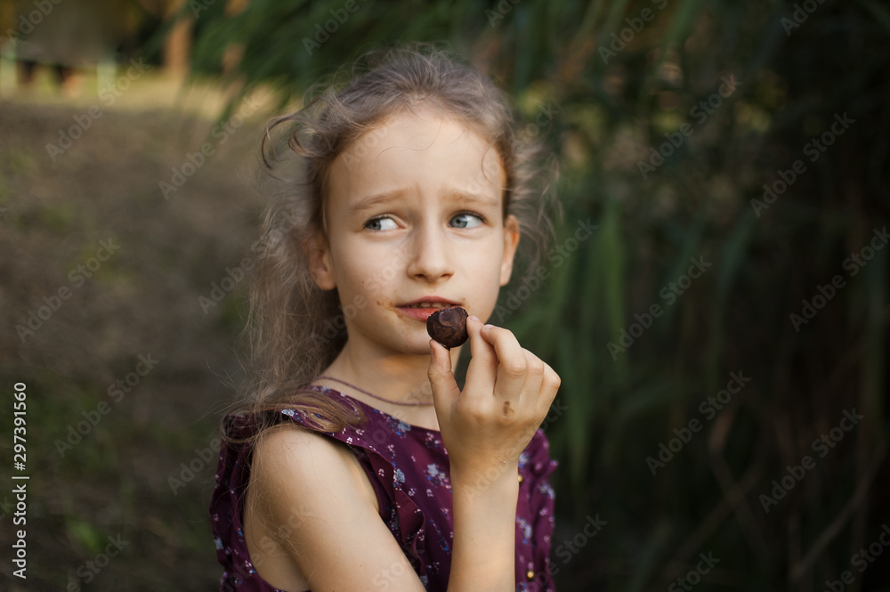Beautiful little blond girl in violet dress with ponytail is eating a chocolate candy in the park durring a day, sugar addiction concept Beautiful little blond girl in violet dress with ponytail is eating a chocolate candy in the park durring a day, sugar addiction concept