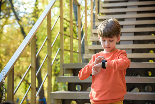 A cute boy wearing stylish shirt stay near school, looking at his smart watch touching the screen. A child using electronic device. Lifestyle concept