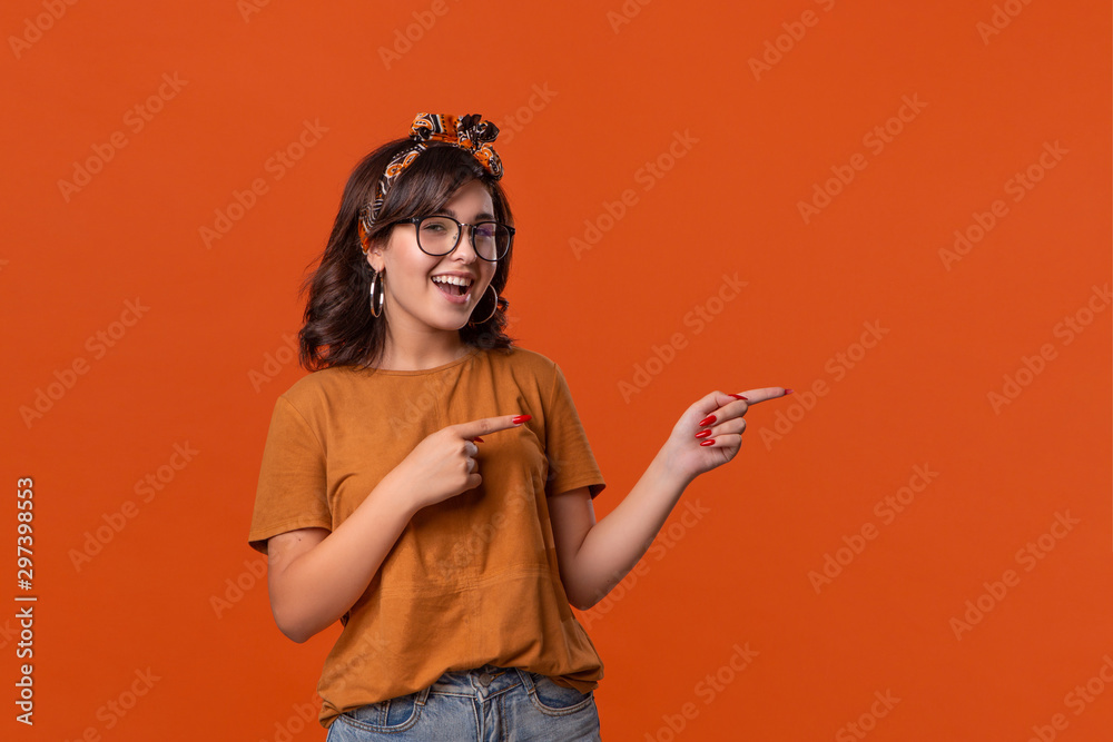 Fototapeta premium Smiling brunette woman in a t-shirt, headband and eyeglasses pointing with her fingers at the right side isolated over orange background.