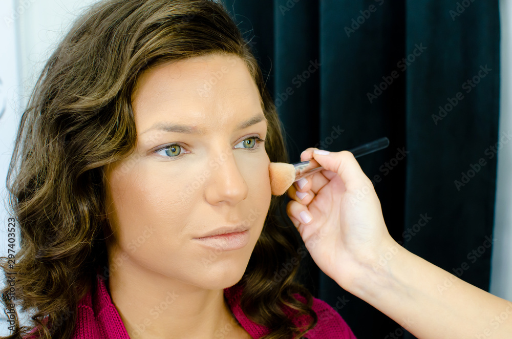 Obraz premium Make-up artist working with brush on model face. Portrait of young woman in beauty studio interior. Close up of Beautiful Young Female Model Putting Blush With Cosmetic Brush
