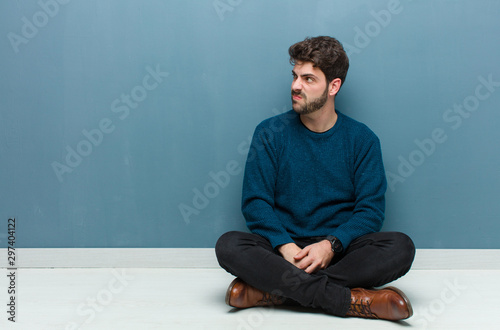 young handsome man sitting on floor open-mouthed in shock and disbelief, with hand on cheek and arm crossed, feeling stupefied and amazed photo