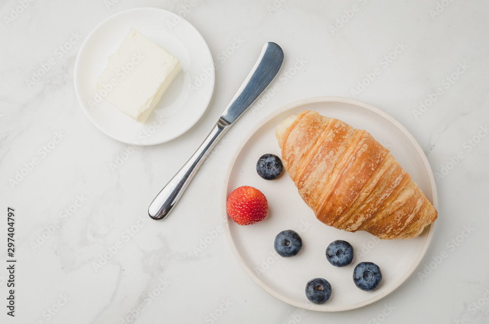 Croissant with berries in white bowl and butter knife on white stone background. Top view