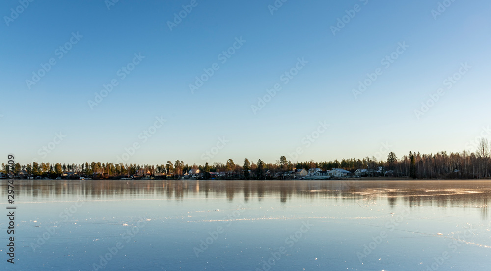 The swedish village lays far away, beside the big crystal clear frozen lake. The surface looks like huge mirror, clear skies, very cold winter day, Northern Sweden