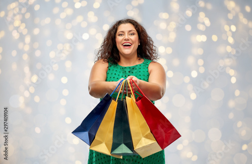 sale, holidays and consumerism concept - happy woman in green dress with shopping bags over festive lights on grey background