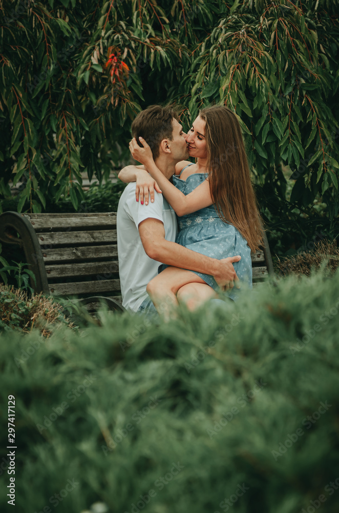 Fototapeta premium Beautiful couple in love sits in a park on a bench in the summer, hugs and kisses. The girl sits on the guy’s lap.