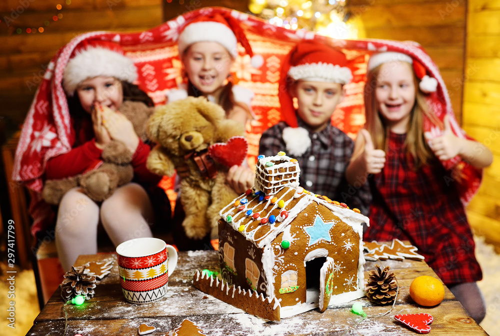 a group of small children friends preschoolers in Santa hats covered with a blanket play with toys and make a gingerbread house on the background of Christmas decor and lights. a group of small children friends preschoolers in Santa hats covered with a blanket play with toys and make a gingerbread house on the background of Christmas decor and lights.