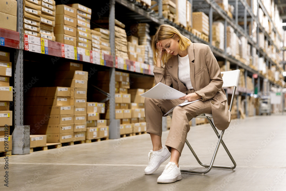 Tired seriously business woman or supervisor in a beige suit looks at documents and holding her forehead, sitting on a chair at empty facility storage/warehouse. Fatigue at work. 