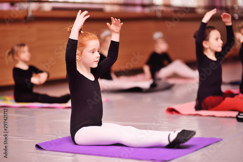 Fototapeta Naklejka Na Ścianę i Meble -  child preschooler - a cute red-haired girl ballerina stretching and doing the splits on the background of ballet schools and groups of children