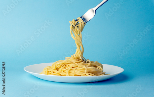 Spaghetti pasta with a fork on a white plate on a blue background. Creative, minimal concept