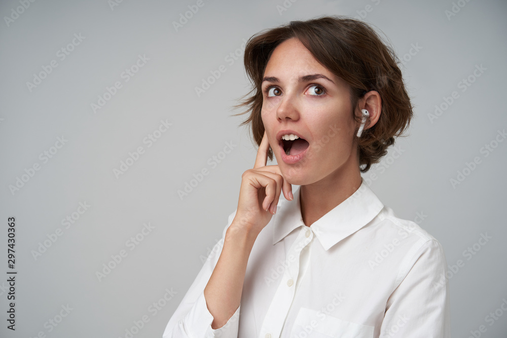 Surprised young beautiful female with short brown hair wearing earphones and looking aside with wide mouth opened, standing over white background in formal clothes Surprised young beautiful female with short brown hair wearing earphones and looking aside with wide mouth opened, standing over white background in formal clothes