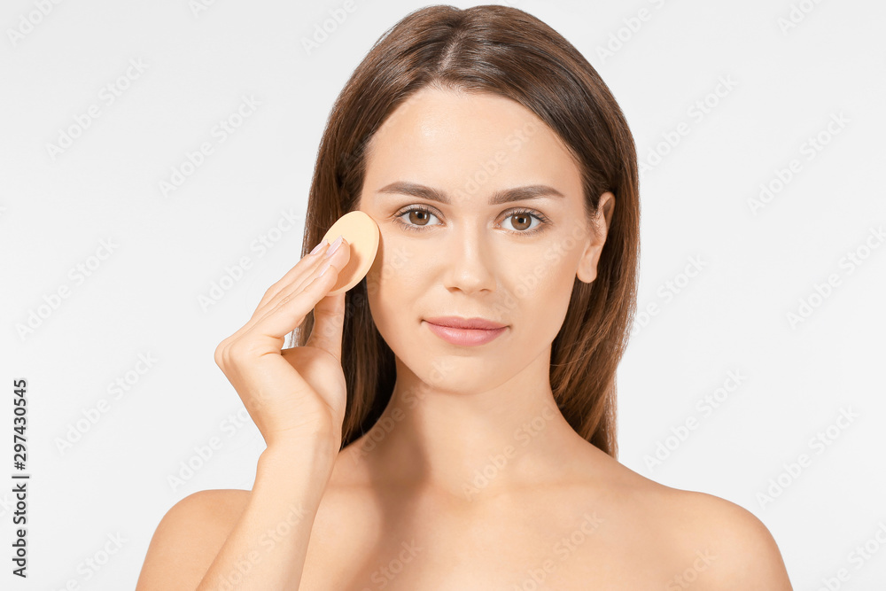 Beautiful young woman applying facial powder against light background