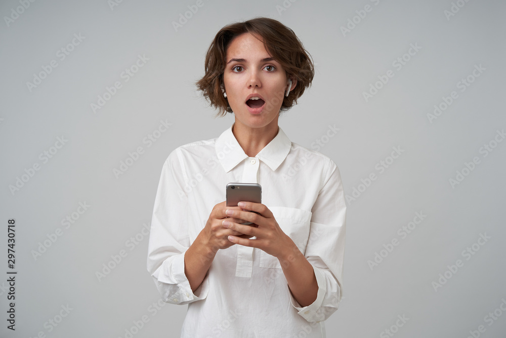 Portrait of surprised pretty brunette female with casual hairstyle holding mobile phone in hands while standing over white background, looking at camera with wide mouth opened Portrait of surprised pretty brunette female with casual hairstyle holding mobile phone in hands while standing over white background, looking at camera with wide mouth opened