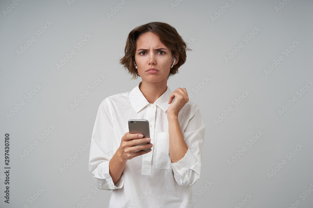 Displeased young brunette woman with casual hairstyle wearing formal clothes while posing over white background, frowning eyebrows and holding smartphone in hand Displeased young brunette woman with casual hairstyle wearing formal clothes while posing over white background, frowning eyebrows and holding smartphone in hand
