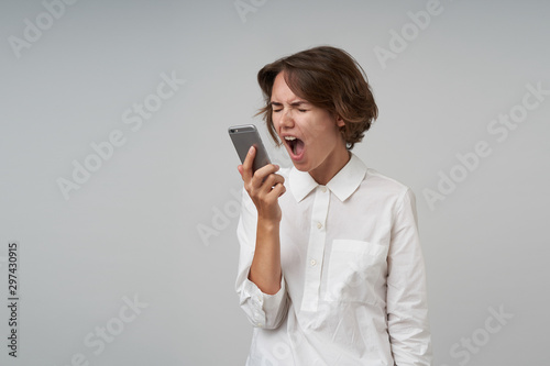 Angry young brunette woman with casual hairstyle shouting to handset loud with wide mouth opened, keeping eyes closed, wearing white shirt while posing over white background