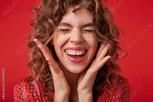 Pleasant looking curly lady with multicolored dots on her face laughing happily and showing her perfect white teeth, keeping eyes closed and raising palms to her face, standing over red background