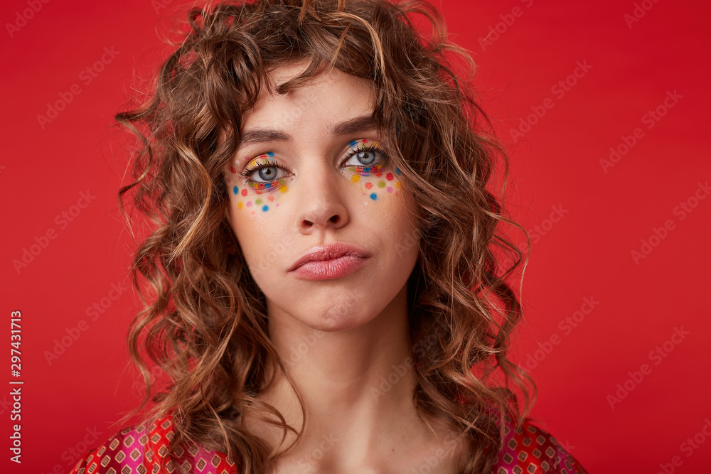 Portrait of beautiful blue-eyed young woman with romantic hairstyle and multicolored dots on her face looking to camera and pursing lips, isolated over red background Portrait of beautiful blue-eyed young woman with romantic hairstyle and multicolored dots on her face looking to camera and pursing lips, isolated over red background
