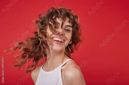 Portrait of happy young pretty woman with curly hair and festive makep laughing cheerfully and looking at camera, posing over red background in white shirt photo