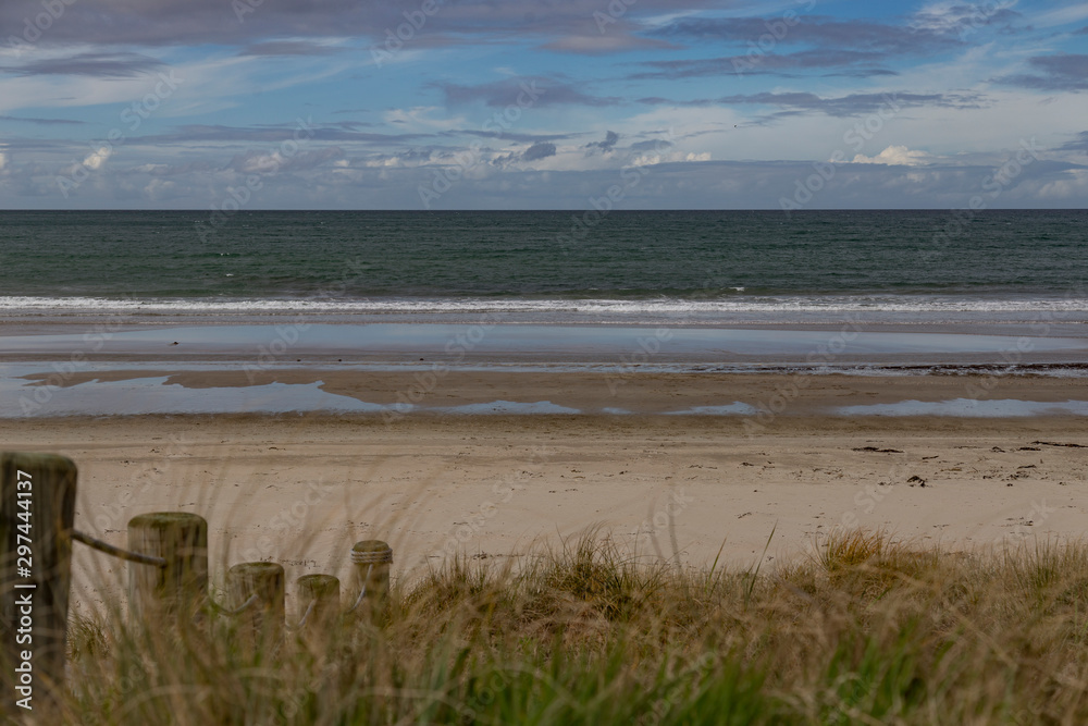 Whangamata, Coromandel Peninsula. View from grass looking straight out to the ocean at low tide. Dune grasses out of focus in the foreground with wooden posts, bollards and rope to protect dunes. Whangamata, Coromandel Peninsula. View from grass looking straight out to the ocean at low tide. Dune grasses out of focus in the foreground with wooden posts, bollards and rope to protect dunes.