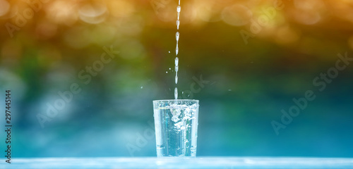 Drink water pouring in to glass over sunlight and natural green background.Water splash in glass Select focus blurred background.