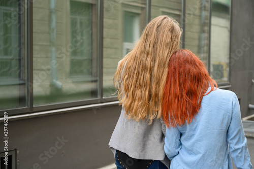 Two friends are walking along the street and gently hugging each other around the waist. The blonde in the jacket and the red-haired woman's dress bowed their heads to each other. View from the back