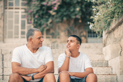 Картината върху платно Teenager son and senior father sitting on stairs outdoors at home, talking