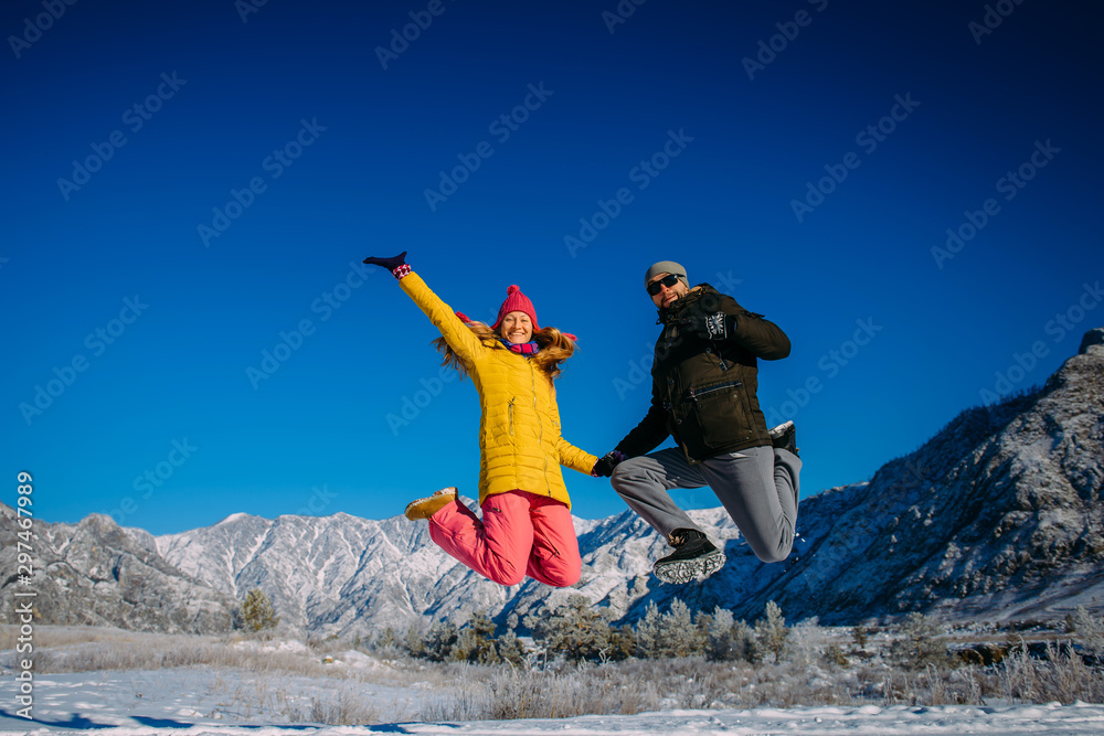 Young cheerful couple in bright clothes jump on the background of snowy mountains on winter sunny day. Guy and girl have fun during new year holidays in the mountains. Christmas and winter activities. Young cheerful couple in bright clothes jump on the background of snowy mountains on winter sunny day. Guy and girl have fun during new year holidays in the mountains. Christmas and winter activities.
