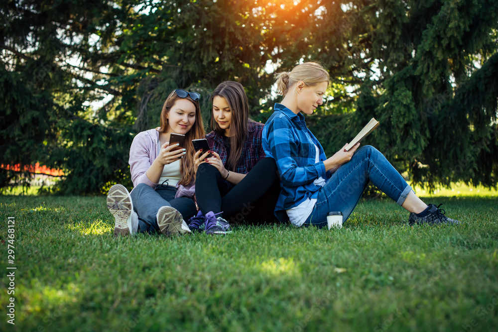 Young girls relax in the park on green grass. Students spend their free time in nature, read books, communicate in social networks. Girlfriends on weekends out of town. Young girls relax in the park on green grass. Students spend their free time in nature, read books, communicate in social networks. Girlfriends on weekends out of town.
