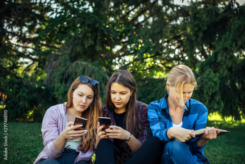 Cute girl students spend free time sitting on green grass in the park on summer day. One woman is reading a book and two young girls are looking at smartphone. Three girlfriends resting on the lawn.