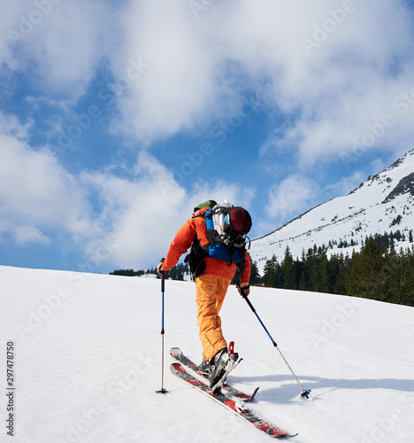 Back view of ski mountaineer walking up along snowy ridge with the skis in the backpack. In background a blue sky. Snow and winter activities, skitouring in mountains.