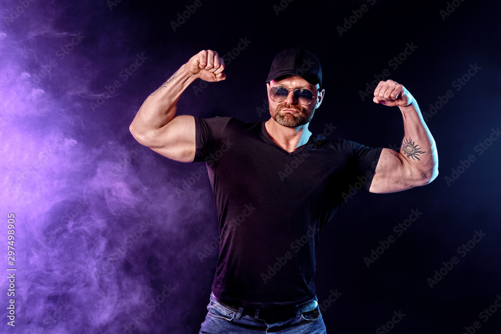 Young strong man bodybuilder in cap on wall background. Dark dramatic colors. Smoke on background.
