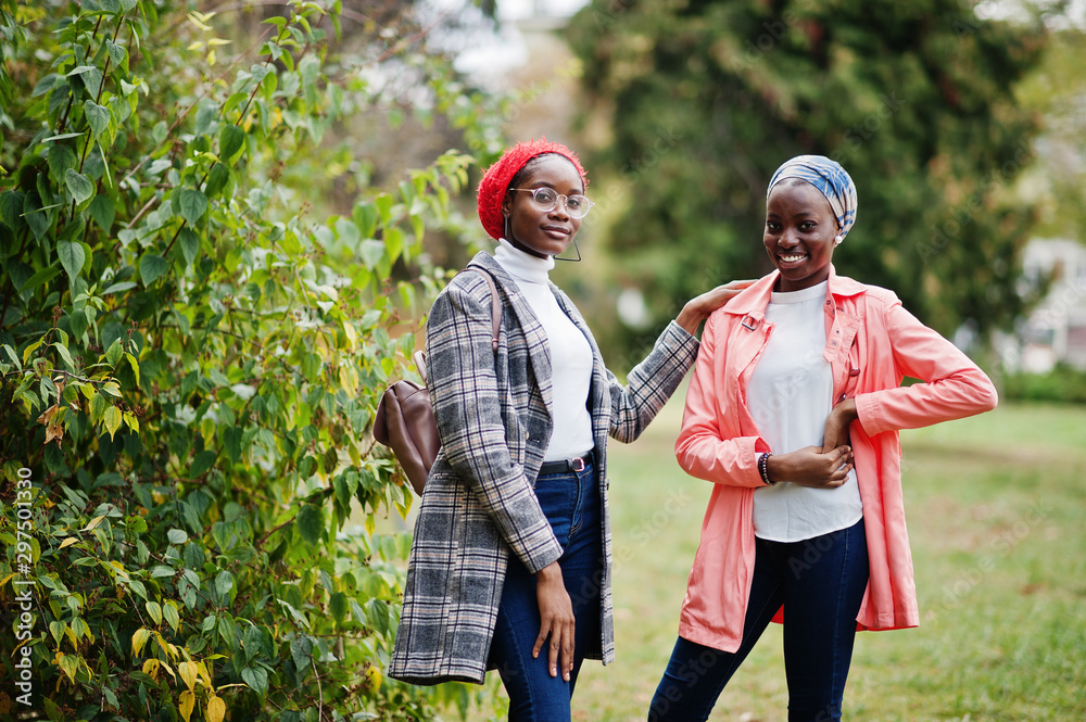 Two young modern fashionable, attractive, tall and slim african muslim womans in hijab or turban head scarf and coat posed at park together.